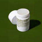 A white jar of Malin+Goetz eucalyptus deodorant with its cap off, lying on its side next to the open jar, set against a solid dark green background.