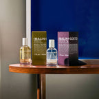 Two MALIN+GOETZ perfume products and their boxes arranged on a wooden table against a split gray and dark blue background. The image includes fig and tobacco eau de parfums.