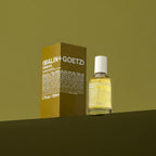 A bottle of (Malin+Goetz) Tobacco eau de parfum with its matching brown cardboard box, displayed on a dark olive green surface against a matching olive green background.